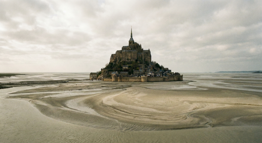 Panoramic view of the Mont Saint-Michel abbey surrounded by sand and tide flats under a cloudy sky. Inside Mont Saint Michel France
