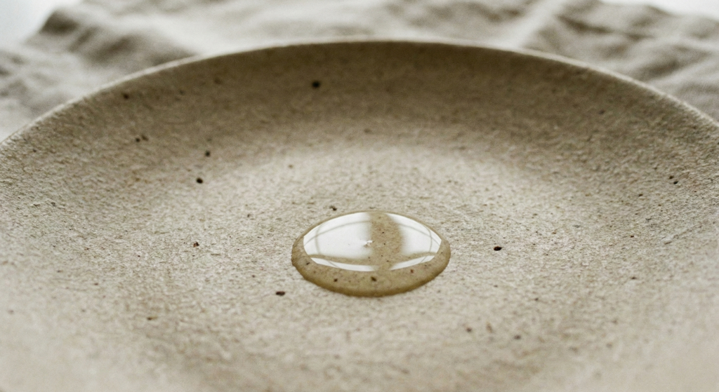 Close-up of a single drop of clear facial oil on a textured beige ceramic plate, representing iconic French skincare.