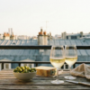 Two glasses of white wine, olives, and cheese on a wooden table with the iconic grey zinc rooftops of Paris in the background.