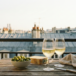 Two glasses of white wine, olives, and cheese on a wooden table with the iconic grey zinc rooftops of Paris in the background.