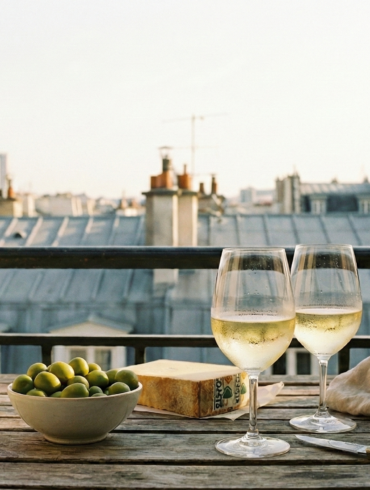 Two glasses of white wine, olives, and cheese on a wooden table with the iconic grey zinc rooftops of Paris in the background.