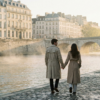 A couple in trench coats holding hands while walking along the Seine River during a misty morning in Paris.