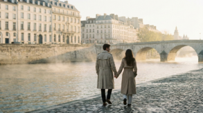 A couple in trench coats holding hands while walking along the Seine River during a misty morning in Paris.
