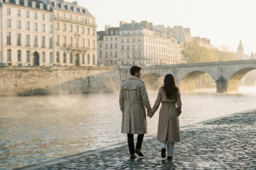 A couple in trench coats holding hands while walking along the Seine River during a misty morning in Paris.
