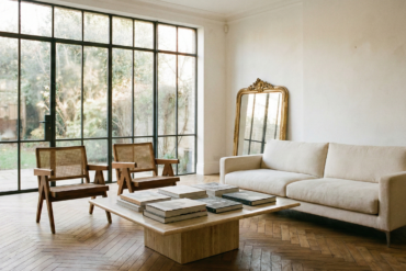 Modern French living room with a neutral sofa, Pierre Jeanneret style chairs, and large windows overlooking a garden.