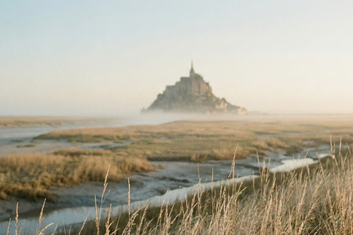 Ethereal view of Mont Saint-Michel emerging from the mist across a marshy landscape at sunrise.