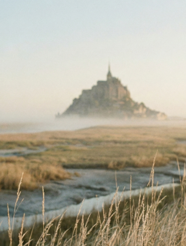 Ethereal view of Mont Saint-Michel emerging from the mist across a marshy landscape at sunrise.