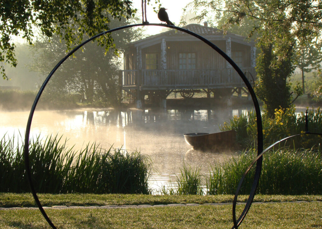 View of a wooden lakeside cabin through a circular metal sculpture by Nicolas Decoster at dawn.