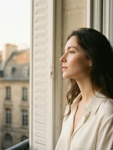 Elegant portrait of a beautiful French woman in an alabaster silk shirt looking out of a Parisian window at golden hour