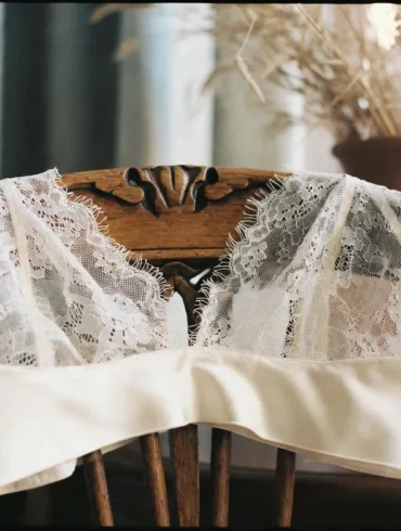 An alabaster silk and Calais lace bralette draped elegantly over a walnut chair in a sun-drenched Parisian apartment