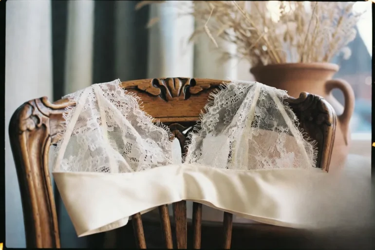 An alabaster silk and Calais lace bralette draped elegantly over a walnut chair in a sun-drenched Parisian apartment