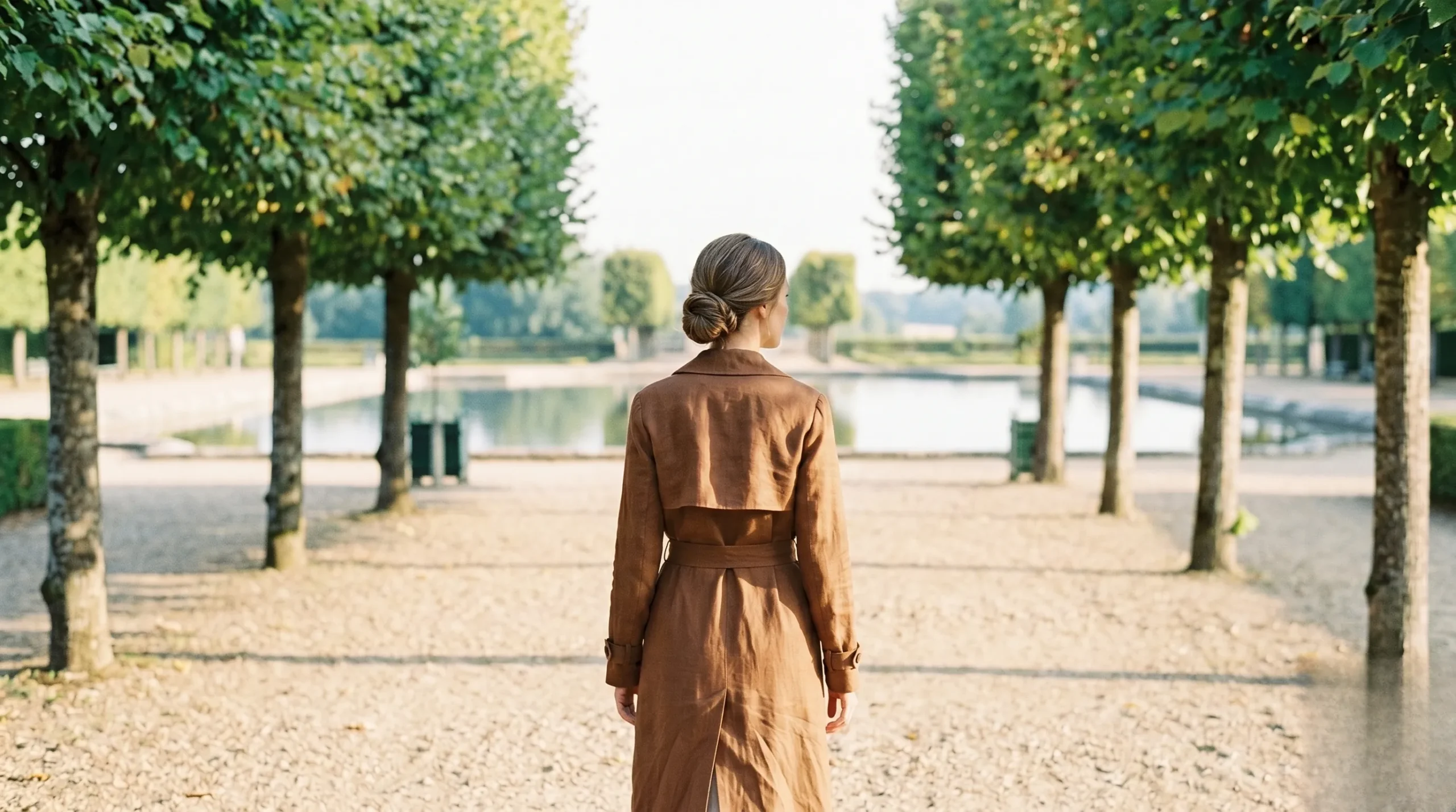 Chic traveler in a trench coat walking along a gravel path next to the serene water mirror at Villandry