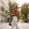 An elegant Parisian woman walking down a sunny street wearing high-waisted alabaster linen trousers and a vibrant terracotta silk shirt