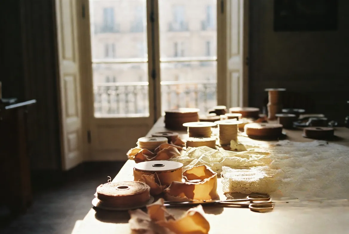 A peaceful Parisian artisan atelier with rolls of sienna silk ribbons, Calais lace, and tailoring tools by a sunlit window