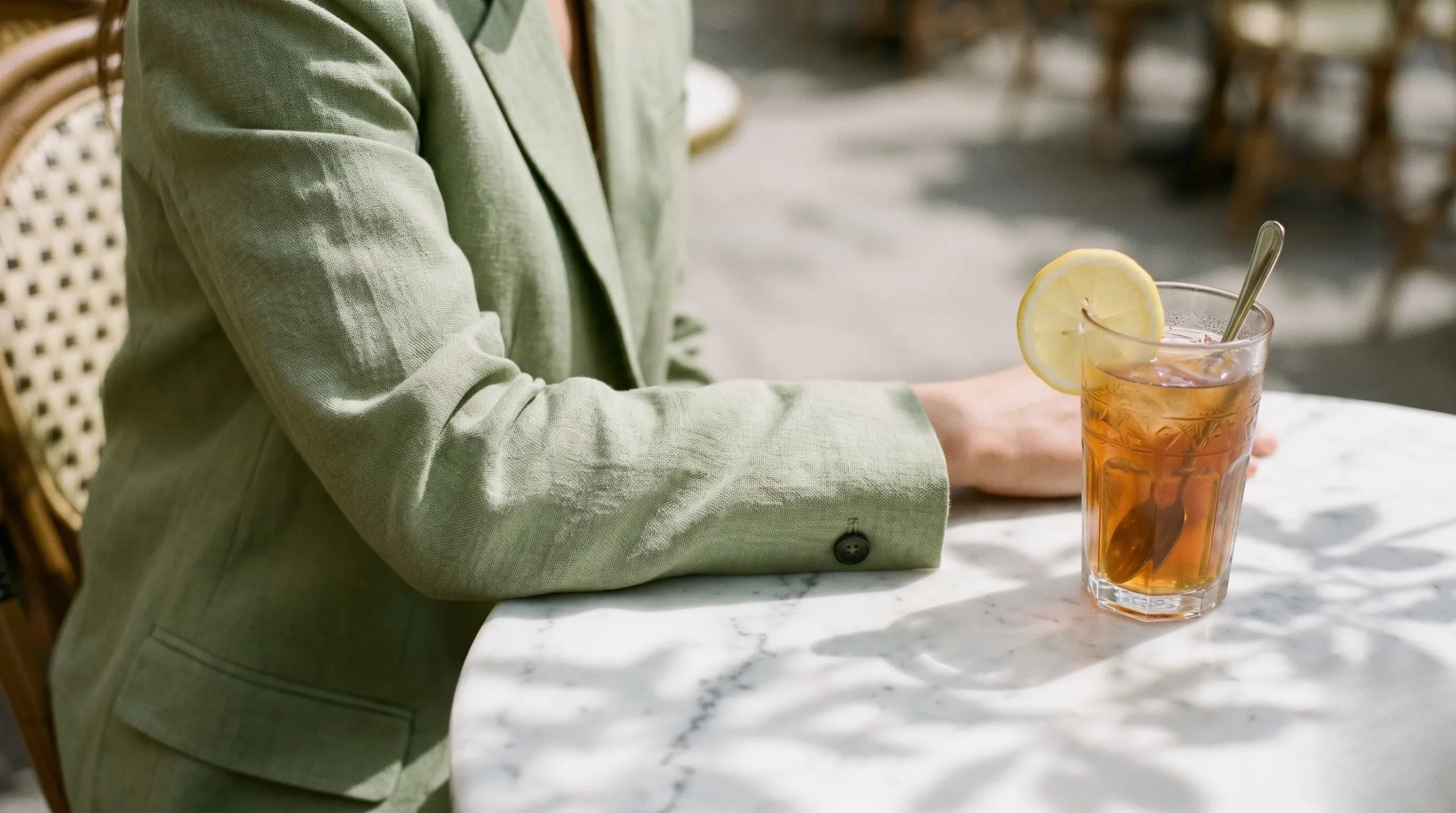 A detailed shot of a woman in a structured pistachio green linen blazer sitting at a marble cafe table with an iced tea