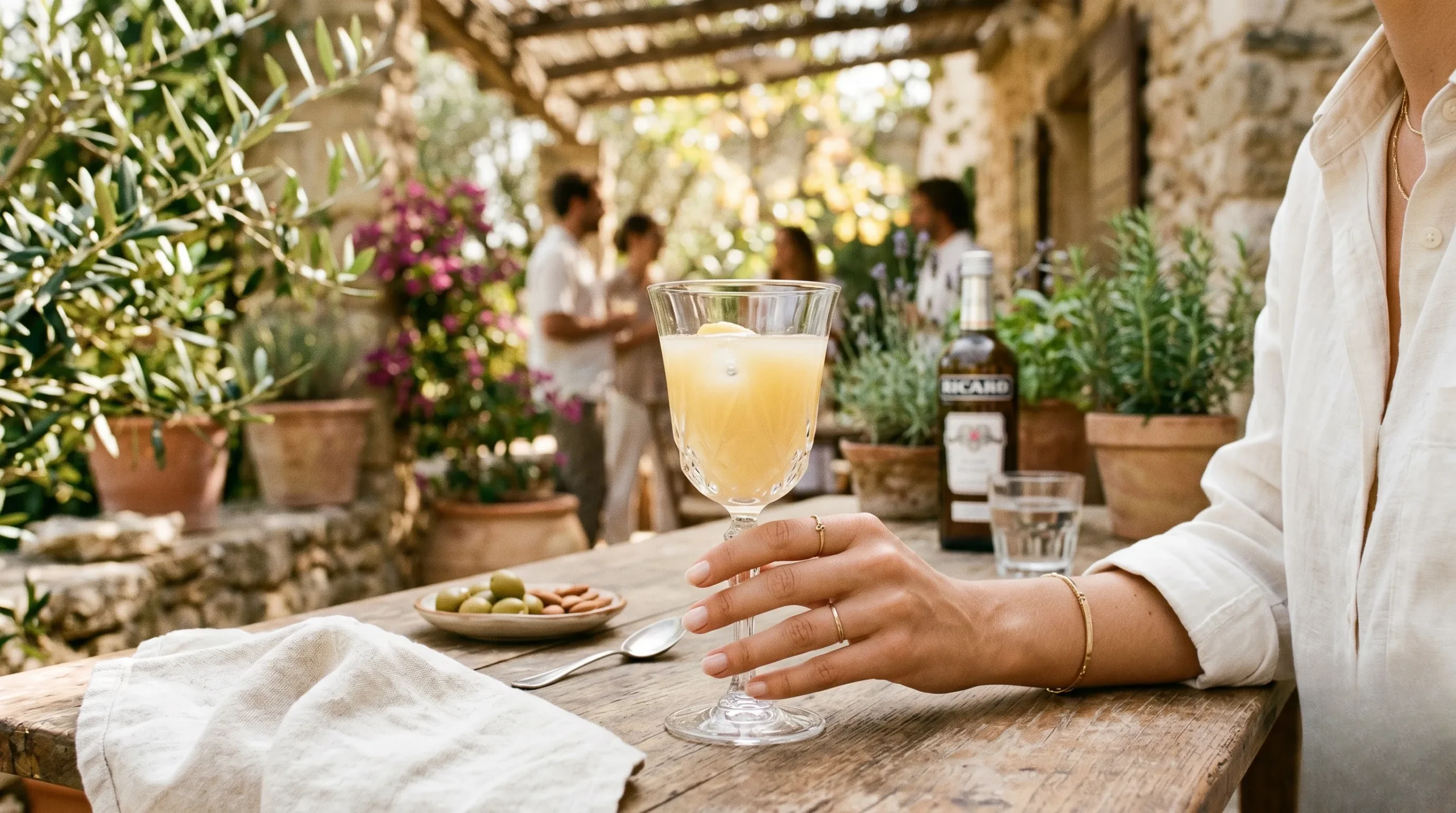 An elegant woman holding a Baccarat glass of cloudy pastis on a Mediterranean outdoor terrace in warm afternoon light. Pastis.