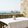 A sun-drenched private stone terrace in Southern France featuring a rustic table with a white linen tablecloth and a ceramic vase of wild lavender overlooking the Mediterranean sea.