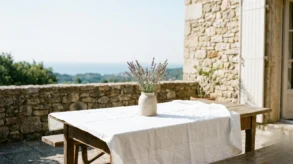 A sun-drenched private stone terrace in Southern France featuring a rustic table with a white linen tablecloth and a ceramic vase of wild lavender overlooking the Mediterranean sea.