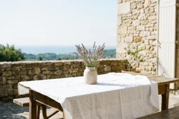 A sun-drenched private stone terrace in Southern France featuring a rustic table with a white linen tablecloth and a ceramic vase of wild lavender overlooking the Mediterranean sea.