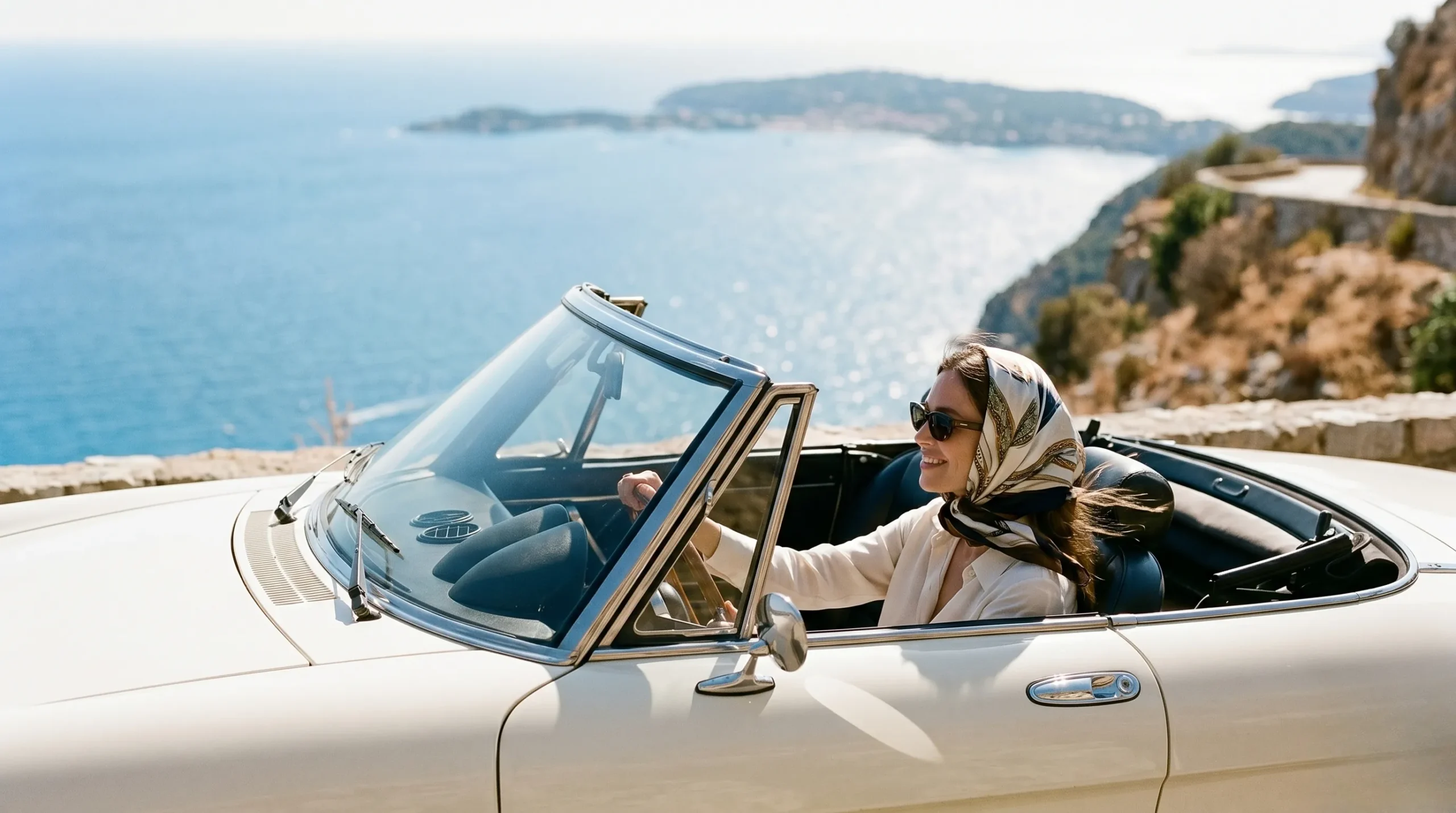 A chic woman wearing a silk headscarf driving a vintage cream Alfa Romeo Spider along a scenic cliff road overlooking the azure French Riviera.
