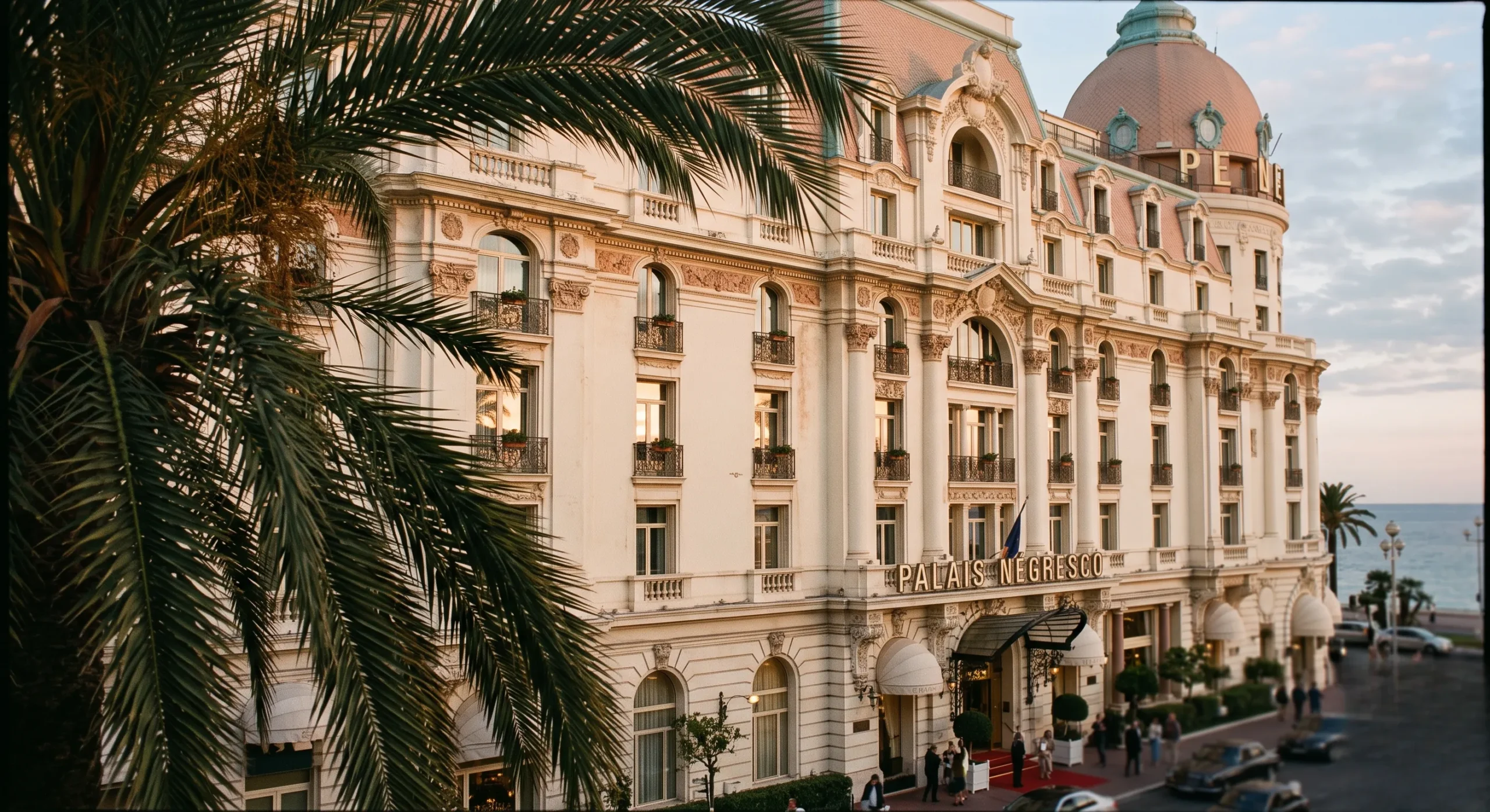 The alabaster exterior facade of a grand Belle Époque hotel in Nice, France, framed by palm leaves under soft sunset light