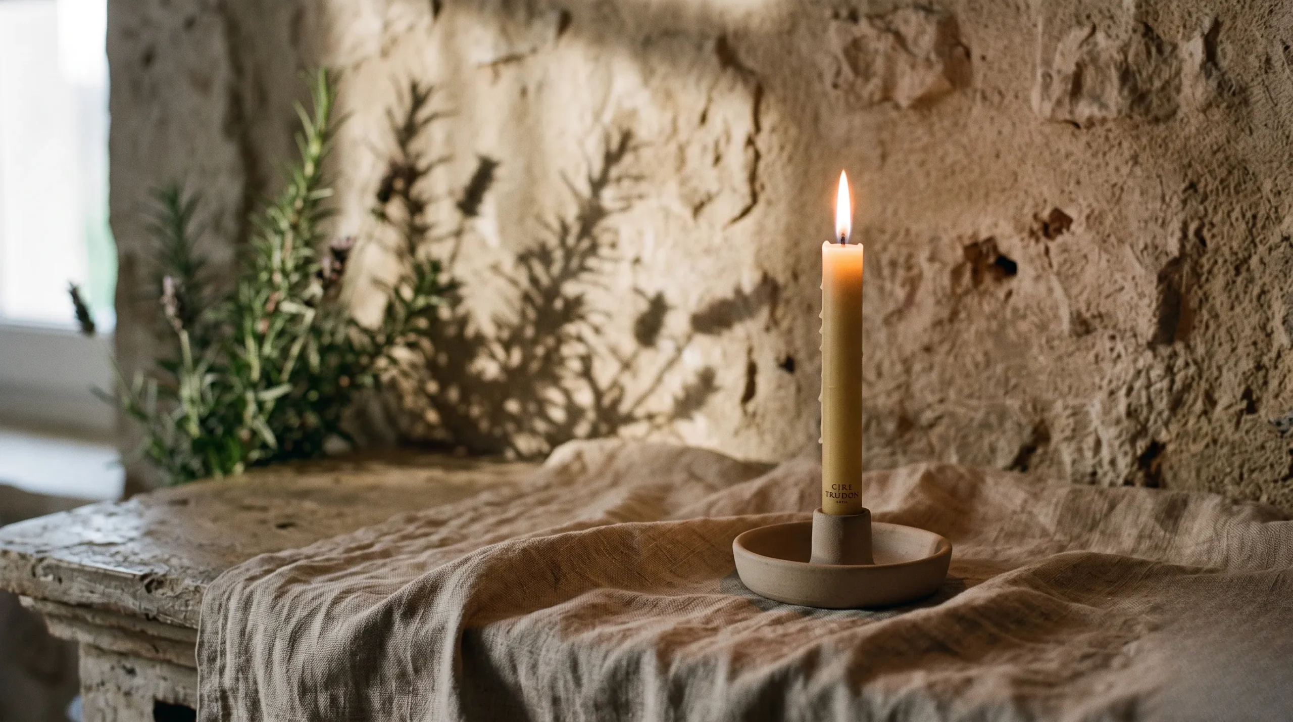 A close-up of a single burning Cire Trudon unscented taper candle casting shadows of rosemary leaves onto a raw draped flax tablecloth.