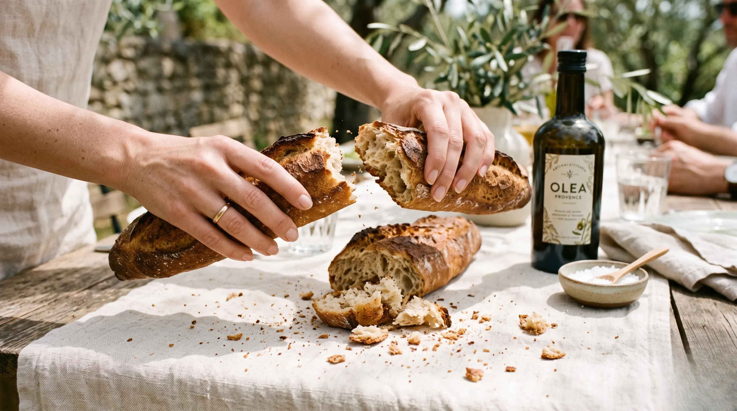 Close-up of elegant hands breaking a crusty rustic baguette over a cream linen tablecloth at an outdoor table in dappled sunlight.