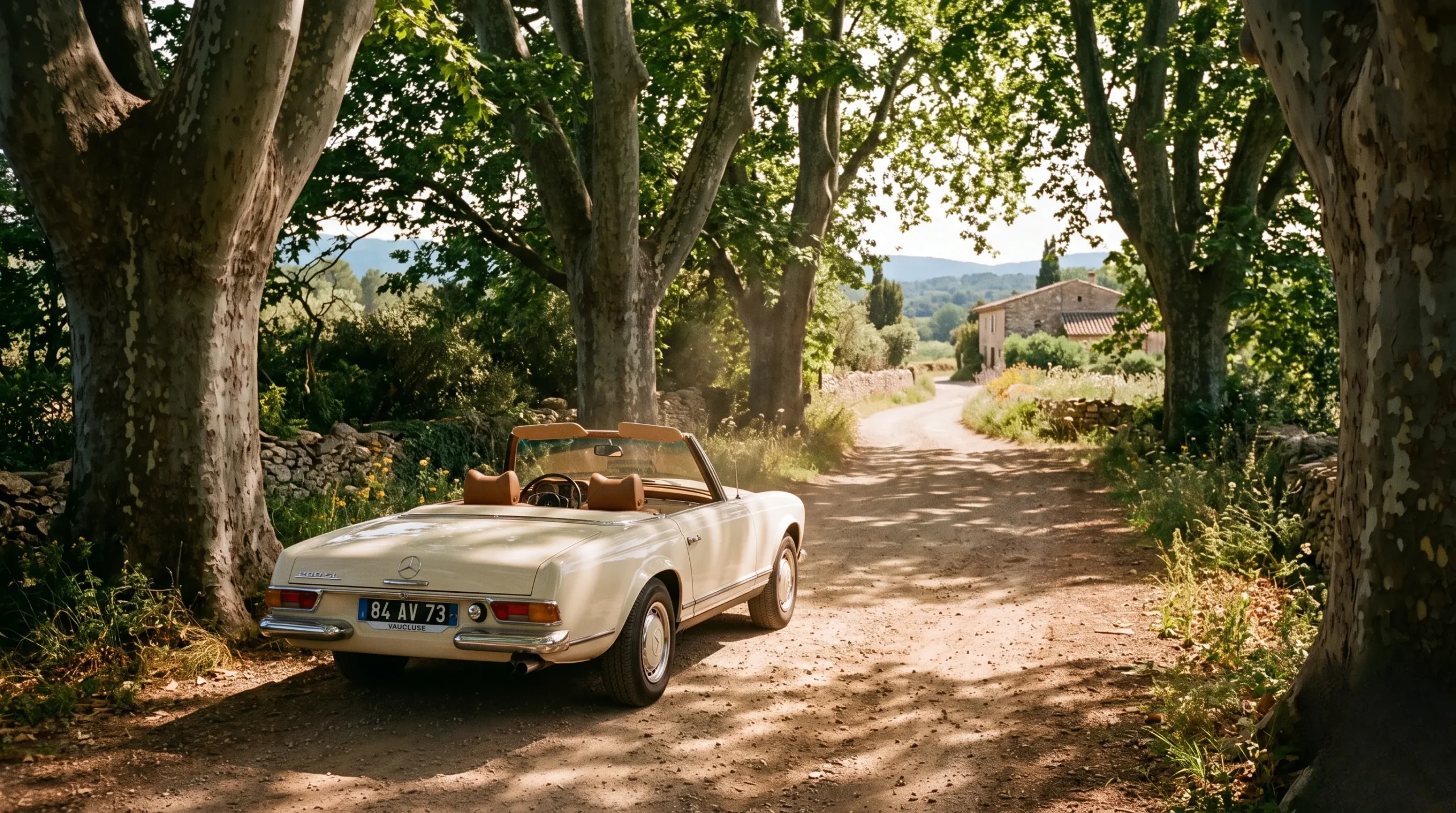 A vintage cream-colored convertible car parked under plane trees on a quiet sienna-toned road in southern France.